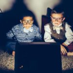 two young boys dressed in business attire and wearing spectacles sit side by side and watch closely an old fashioned television box receiver with built in wire ariels.