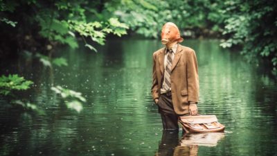 A man in a suit and holding a briefcase with a fish-head stands knee-deep in water.