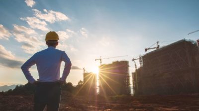 A man stands with hands on hips surveying construction of three high-rise buildings.