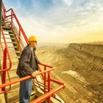 An engineer takes a break on a staircase and looks out over a huge open pit coal mine as the sun rises in the background.