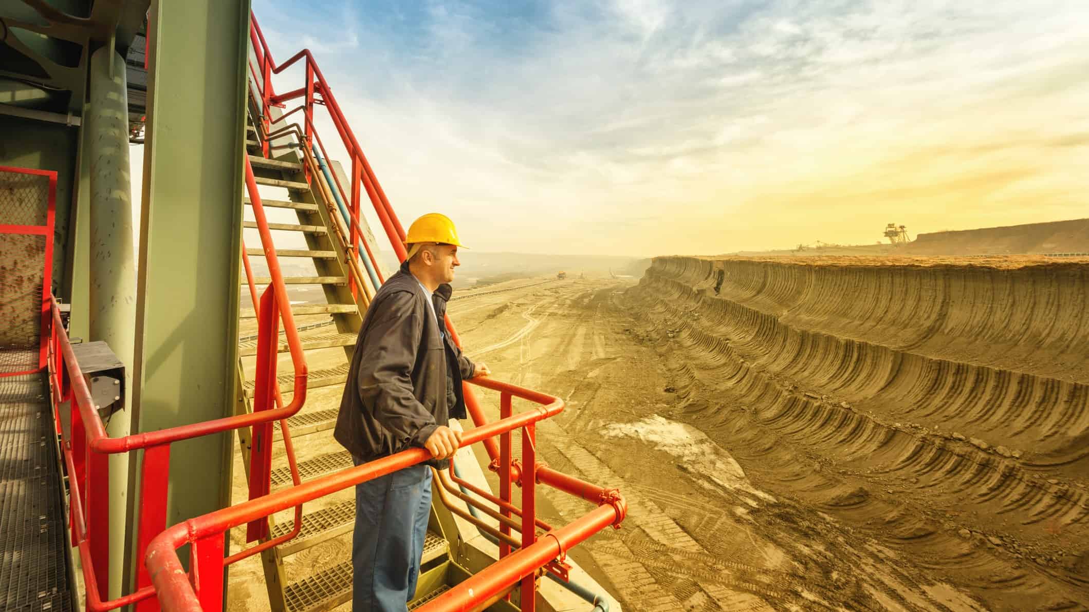 An engineer takes a break on a staircase and looks out over a huge open pit coal mine as the sun rises in the background