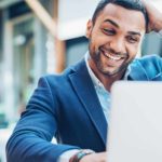 An executive in a suit smooths his hair and laughs as he looks at his laptop feeling surprised and delighted.