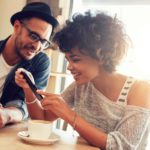 A happy man and woman sit having a coffee in a cafe while she holds up her phone to show him the ASX shares that did best today.