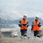 Two mining workers in orange high vis vests walk and talk at a mining site