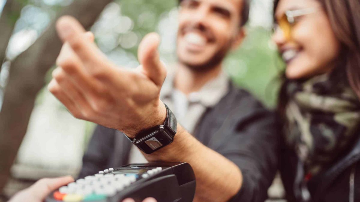 A laughing man standing next to a woman holds out his arm to a payments machine to pay with his smartwatch