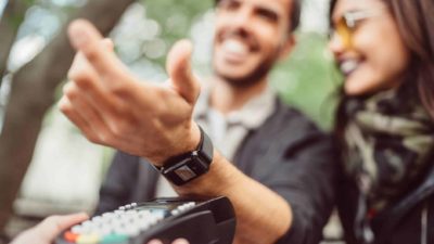 A laughing man standing next to a woman holds out his arm to a payments machine to pay with his smartwatch