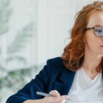 A female sharemarket analyst with red hair and wearing glasses looks at her computer screen watching share price movements.