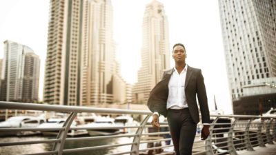 A well-dressed man strides along a river bank with large buildings behind.