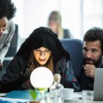 A fortune teller looks into a crystal ball in an office surrounded by business people.