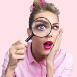 A female ASX investor looks through a magnifying glass that enlarges her eye and holds her hand to her face with her mouth open as if looking at something of great interest or surprise.