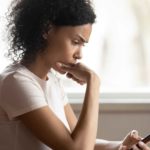 a woman looks down at her phone with a look of concern on her face and her hand held to her chin while she seriously digests the news she is receiving.