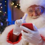 santa looks intently at his mobile phone with gloved finger raised and christmas tree in the background.