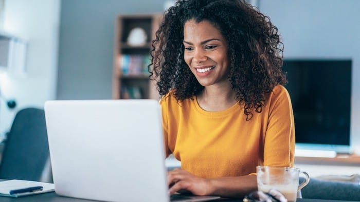 a smiling woman sits at her computer at home with a coffee alongside her, as if pleased with her investments.