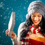 A young woman wearing a beanie as the snow falls around her smiles and opens a Christmas present in a box looking excited and smiling to represent the special dividend for Grange Resources shareholders announced today