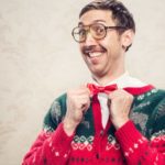 a nerdish looking man with a lovely big smile adjusts his bow tie and raises his eyebrows behind his thick glasses, as he wears a heavy knitted Christmas sweater in traditional Christmas colours of green and red.