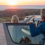 A couple hang off their car looking at the sun rising over the horizon.