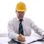 A man wearing a shirt, tie and hard hat sits in an office and marks dates in his diary.