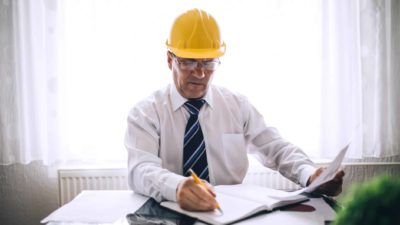 A man wearing a shirt, tie and hard hat sits in an office and marks dates in his diary.