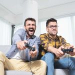 Two men sit side by side on a couch with video game controls in their hands and expressive looks on their faces as they react to the action in front of them in a home setting.