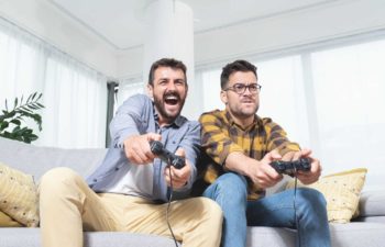 Two men sit side by side on a couch with video game controls in their hands and expressive looks on their faces as they react to the action in front of them in a home setting.