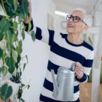 a mature but cool older woman holds a watering can and tends to a healthy green plant growing up the wall in her house.