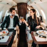 a couple clink champagne glasses on board a private aircraft with gourmet food plates set in front of them. They are wearing designer clothes and looking wealthy.