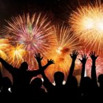 a group of people in shadow profile leap and hold their arms high in wonder of a fireworks display that fills the sky with light and colour and spectacular shapes.