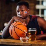 a man sits at a bar leaning sadly on his basketball wearing a US flag sticker on his cheekbone near a half drunk beer and looking despondent as though his basketball team has just lost a game.