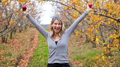 a girl stands in an apple orchard holding two red apples in raised arms with a happy, celebratory look on her face with a large smile and a pretty country background to the picture.