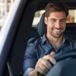 A handsome smiling man sits in the front seat of an electric vehicle with his hands on the wheel feeling pleased that the Carsales share price is going up and the company will shortly pay its biggest dividend ever