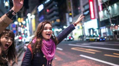 Two women hold their hands up in the street with electric billboards shining brightly behind them.