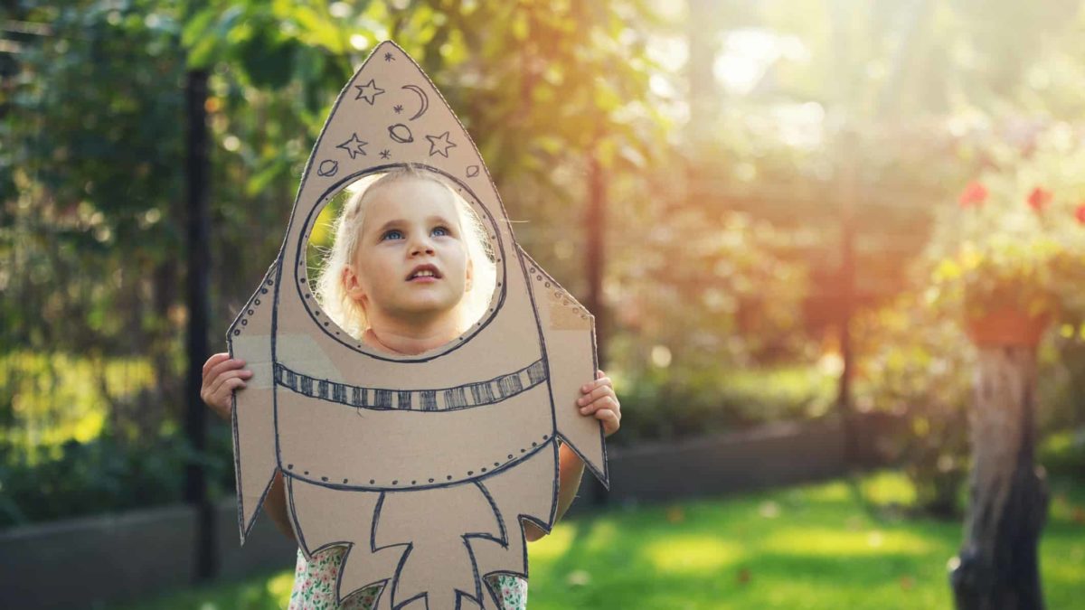 A little girls looks up longingly through a rocket she has made from cardboard, dreaming of shooting to the stars one day.