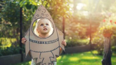 A little girls looks up longingly through a rocket she has made from cardboard, dreaming of shooting to the stars one day.