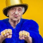 An older female ASX investor holds a gangster-style fist pump pose showing off gold rings with dollar signs on them.