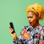 A woman with bright yellow hair wearing a brightly patterned blouse reacts to big news that she's reading on her phone.