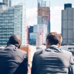 Two businessmen look out at the city from the top of a tall building.