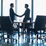 A silhouette shot of two business man shake hands in a boardroom setting with light coming from full length glass windows beyond them.