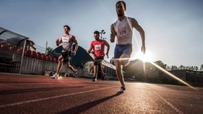 Three male athletes sprint on an athletics track with the sun low on the horizon behind them representing the race between ASX lithium shares to outperform