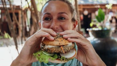 A woman smiles as she holds up a vegan burger to her mouth.