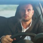 a car driver sits up and looks alert with wide eyes and an expression of concentration while he holds the wheel of a car.