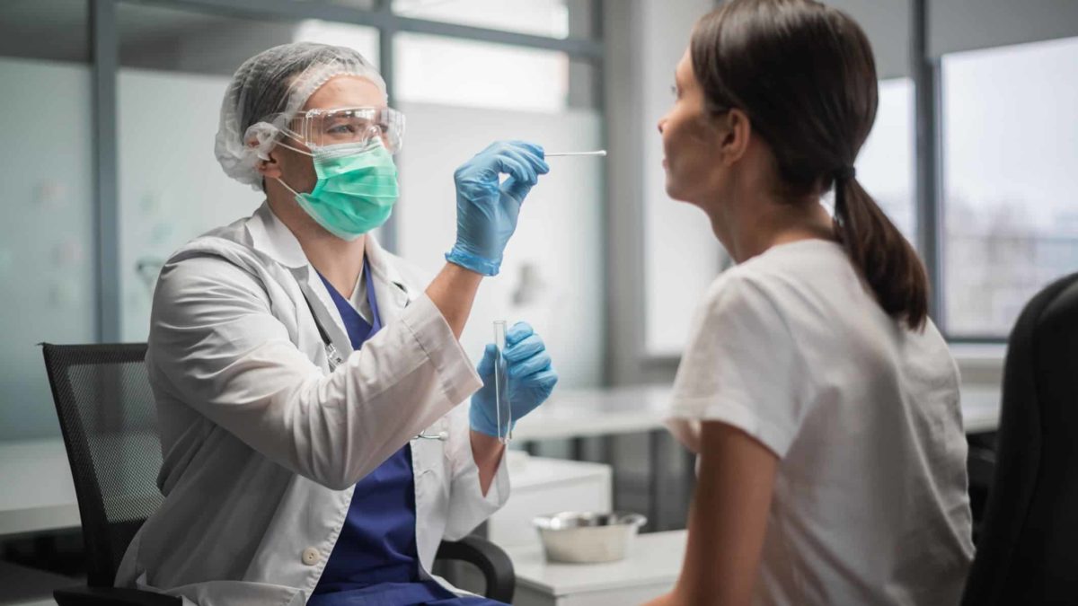 a medical person in full protective gear with gloves and goggles administers a swab to a young woman's nose in a COVID-19 PRC test.