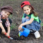 two children squat down in the dirt with gardening tools and a watering can wearing denim overalls and smiling very sweetly.