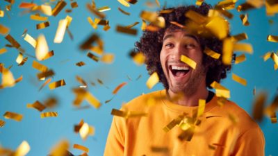 a man wearing a gold shirt smiles widely as he is engulfed in a shower of gold confetti falling from the sky. representing a new gold discovery by ASX mining share OzAurum Resources