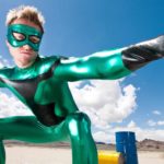 a man dressed in a green superhero lycra outfit stands in a crouched pose with arms outstretched as if ready to spring into action with a blue sky and oil barrels lying in the background.