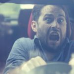 a wide-mouthed man looks scared as he grips the wheel of a car while driving in a murky environment.