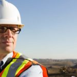 A man wearing a hard hat and high visibility vest looks out over a vast plain where heavy mining equipment can be seen in the background.