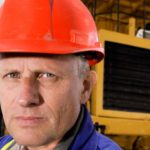 A man wearing a hard hat stands in front of heavy mining machinery with a serious look on his face.