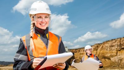 A female miner wearing a high vis vest and hard hard smiles and holds a clipboard while inspecting a mine site with a colleague.