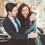 A young couple hug each other and smile at the camera standing in front of their brand new luxury car