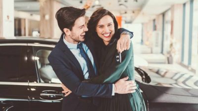 A young couple hug each other and smile at the camera standing in front of their brand new luxury car